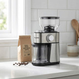 Silver electric coffee grinder with bean container on kitchen counter next to coffee beans and a bag.