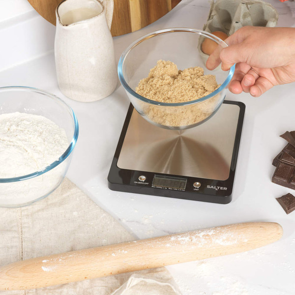 Black digital kitchen scale with stainless steel platform, shown for baking with ingredients.
