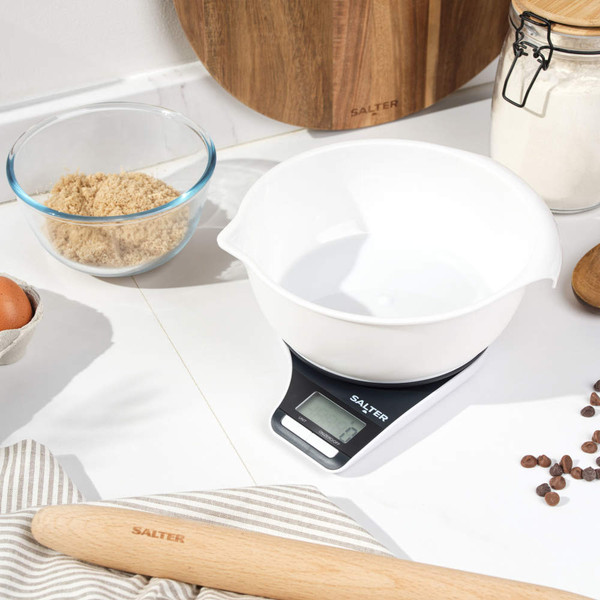 Digital kitchen scale with white mixing bowl on worktop, surrounded by baking ingredients
