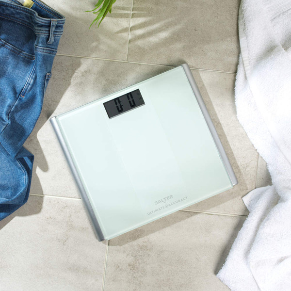 Person wearing beige and white trainers stands on a glass bathroom scale, light tile floor.
