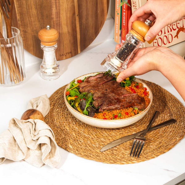 Hand grinding black pepper onto steak salad in bowl, for kitchenware product listing.