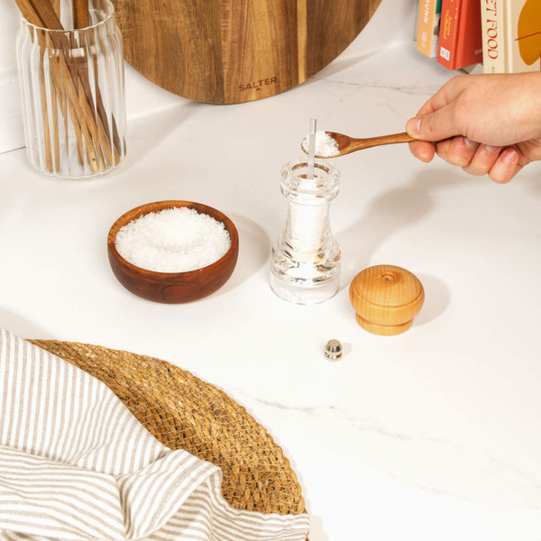 Clear pepper mill with wooden lid being refilled with black peppercorns on a kitchen counter