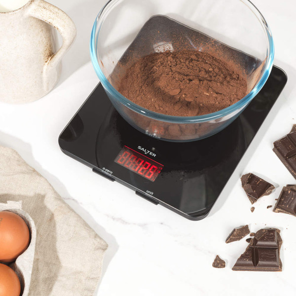 Black digital kitchen scale with red LED display on worktop, surrounded by chocolate pieces
