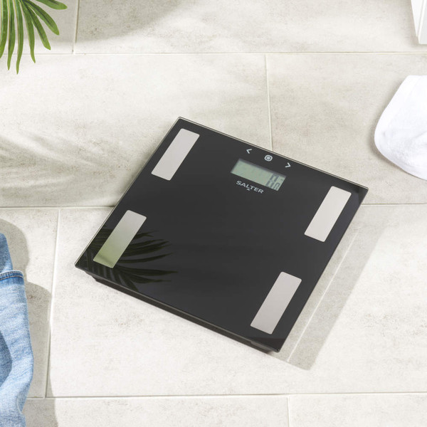 Black digital bathroom scale with silver sensors, top-down view on grey tiles.