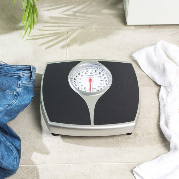 Mechanical bathroom scale with black platform on tiled floor, jeans and towel nearby