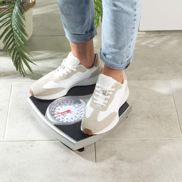 Mechanical bathroom scale with black platform on tiled floor, jeans and towel nearby