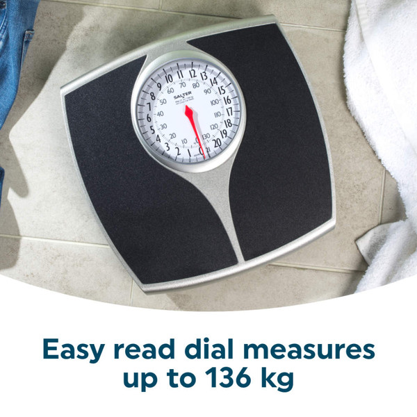 Mechanical bathroom scale with black platform on tiled floor, jeans and towel nearby