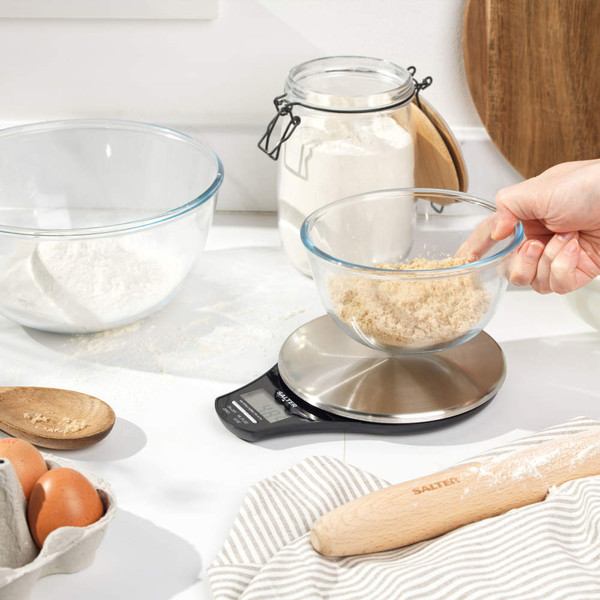 Kitchen scene with hand weighing brown sugar in glass bowl on digital Salter scale