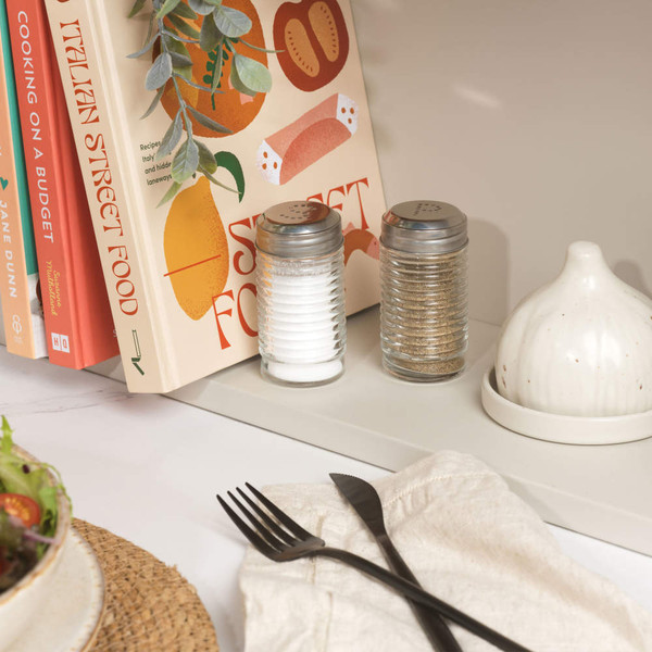 Glass salt and pepper shakers on kitchen shelf beside cookbooks and ceramic butter dish.