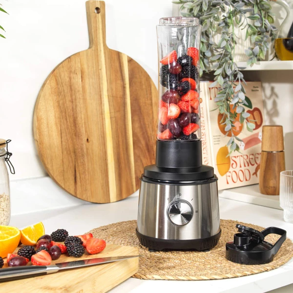 Silver blender with jug filled with fresh fruit, travel bottle, and assorted fruit beside