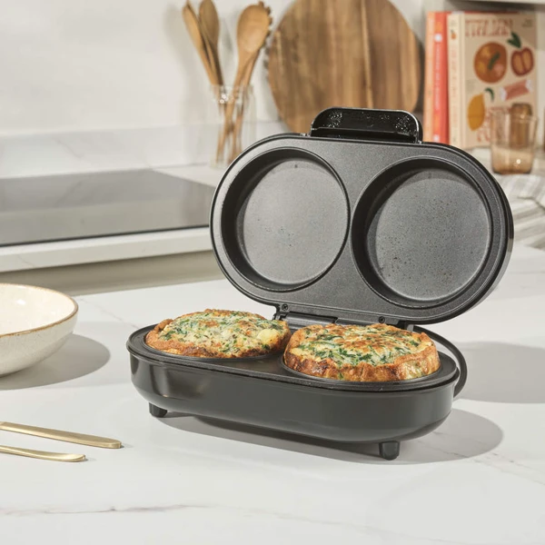 Black electric omelette maker open with two cooked omelettes inside, shown on kitchen counter