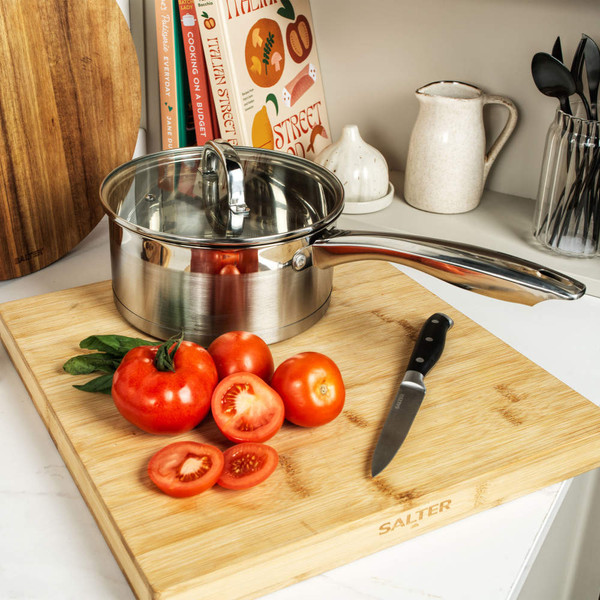 Stainless steel saucepan with lid on wooden chopping board beside fresh tomatoes and knife