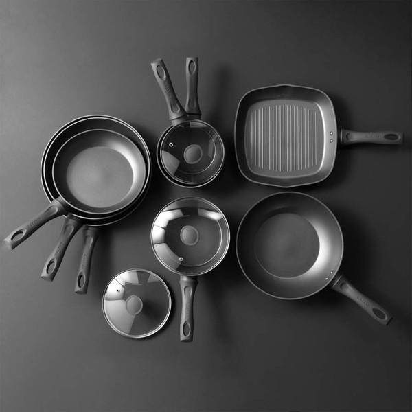 Set of non-stick black saucepans, frying pans and griddle pan with glass lids, arranged on grey background