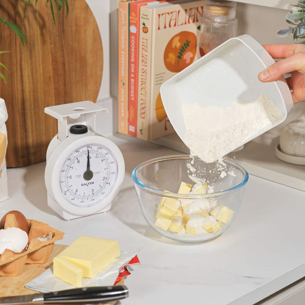 Kitchen scene with white mechanical scales, butter, eggs and flour being poured into a glass bowl