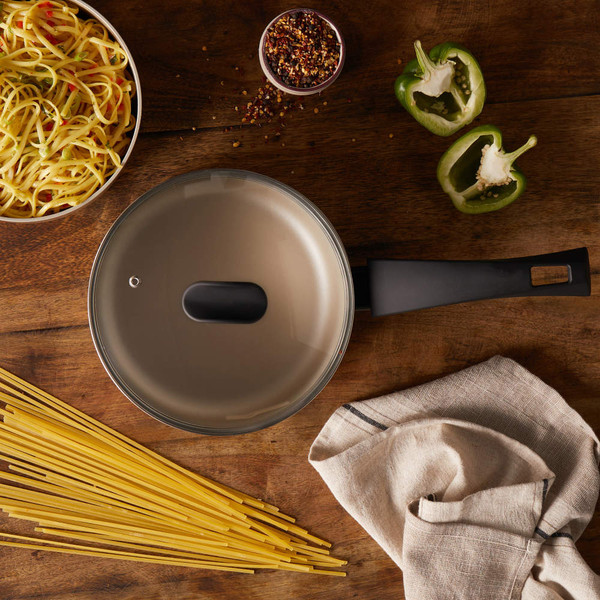 Non-stick saucepan with lid and black handle on wooden surface with spaghetti and vegetables.