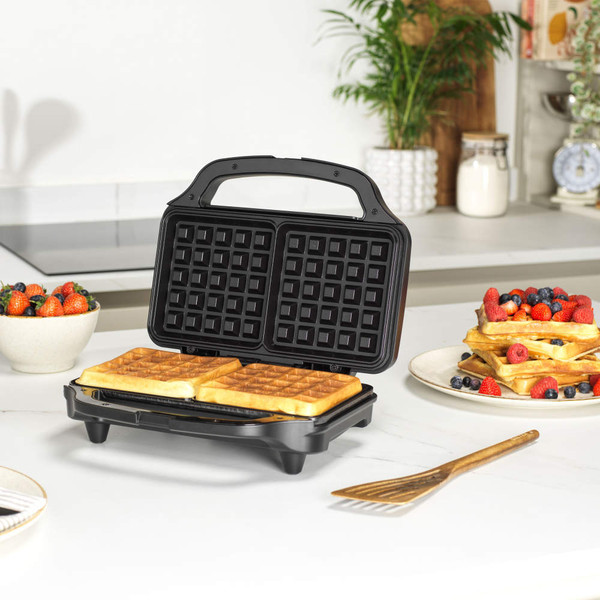 Black waffle maker with two square waffles cooking, shown on kitchen counter.