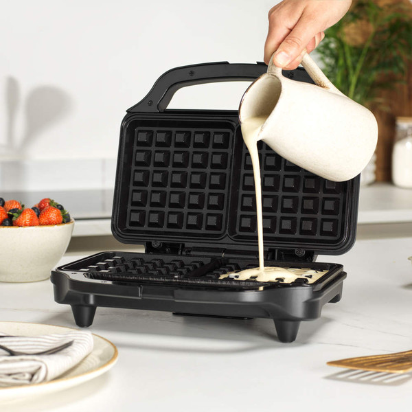 Waffle maker open with batter being poured in, shown on kitchen counter.