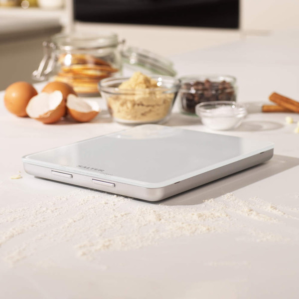 Digital kitchen scale with butter in bowl, baking ingredients and egg on white surface