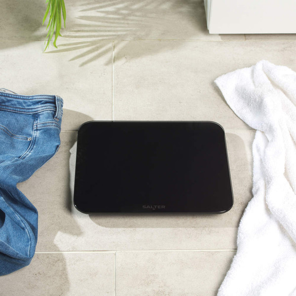 Black glass bathroom scale on tiled floor with jeans and white towel beside