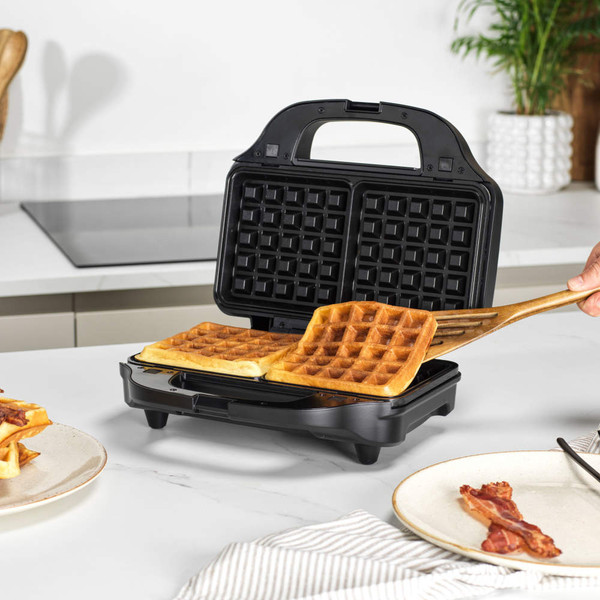 Black sandwich and waffle maker with interchangeable plates, shown making two golden waffles on a kitchen worktop.
