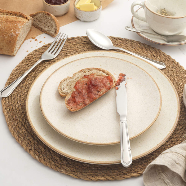 Table setting with seeded bread and strawberry jam on cream stoneware plates, cutlery shown.