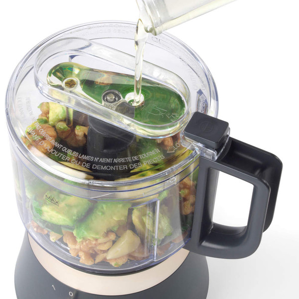 Food processor with avocado, walnuts, and garlic, oil being poured in through lid