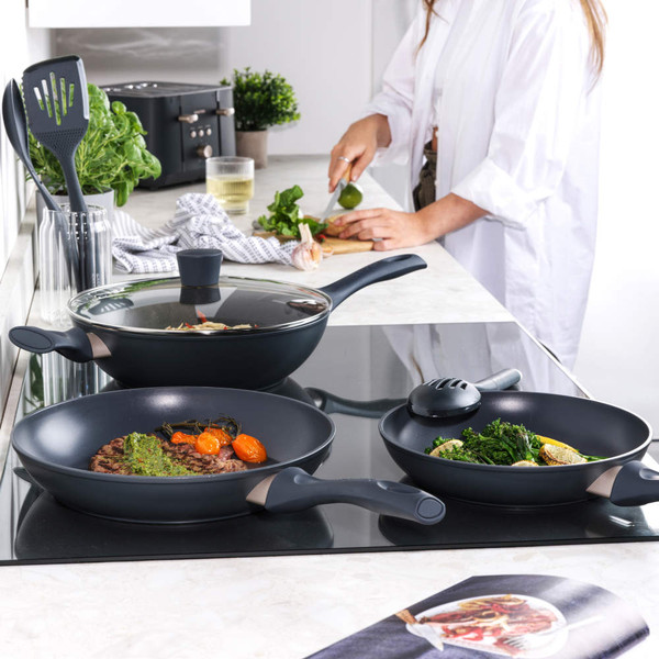 Set of three non-stick black frying pans on hob with food, person preparing ingredients in background