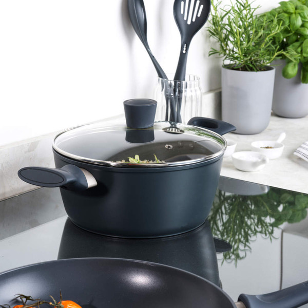 Dark blue non-stick saucepan with glass lid on induction hob, kitchen utensils and herbs in background