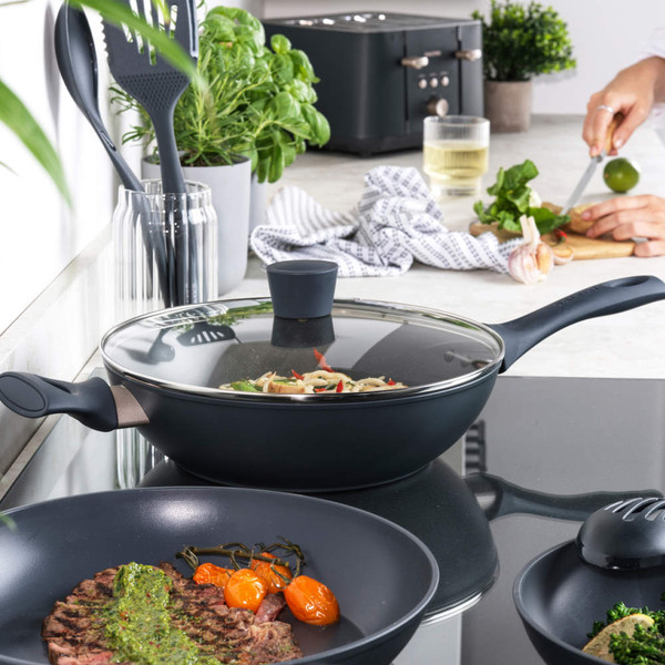 Non-stick frying pan set with glass lid on hob, herbs and utensils in modern kitchen