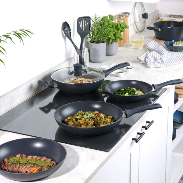 Set of black non-stick frying pans and wok on hob, some with cooked food, in modern kitchen