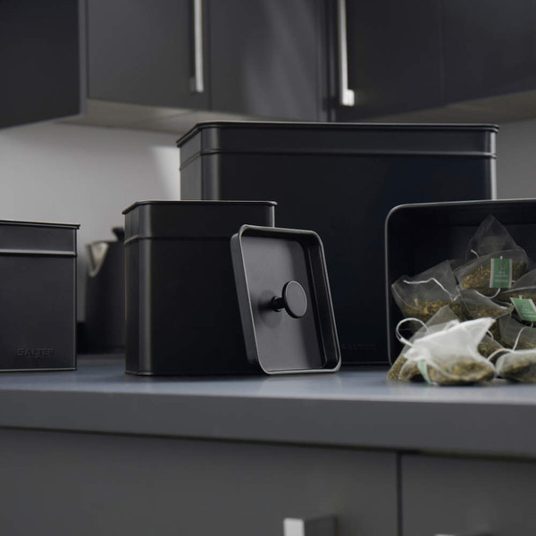 Set of black metal kitchen storage tins, one open showing tea bags inside.