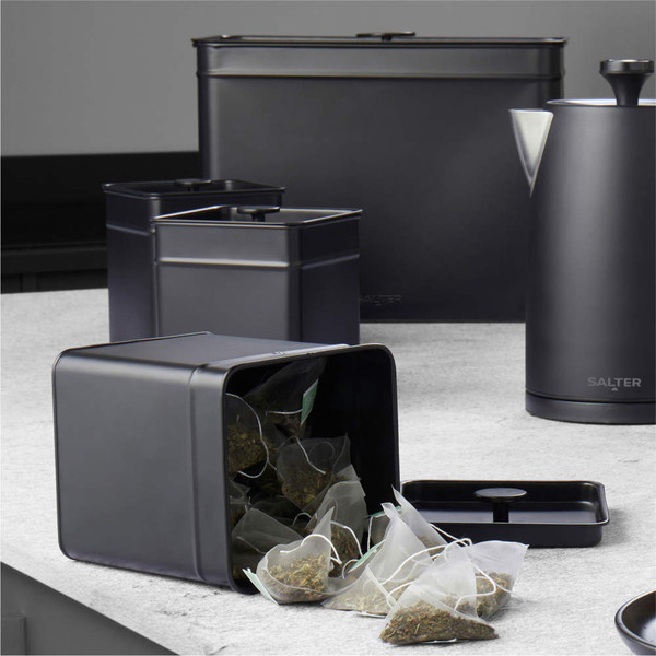 Black metal tea caddy with lid removed, loose tea bags visible; matching kitchen canisters and kettle.