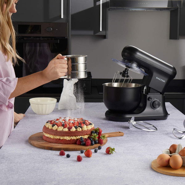 Black stand mixer on kitchen counter with cake, eggs, and baking tools.