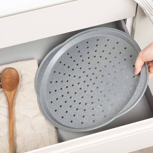 Grey perforated pizza tray shown in kitchen drawer, hand placing tray inside.