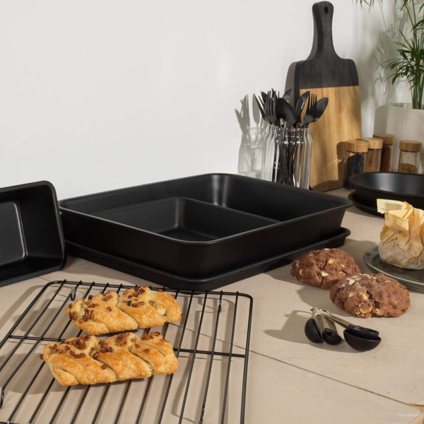 Set of black non-stick baking trays on kitchen counter with cooling rack and pastries.