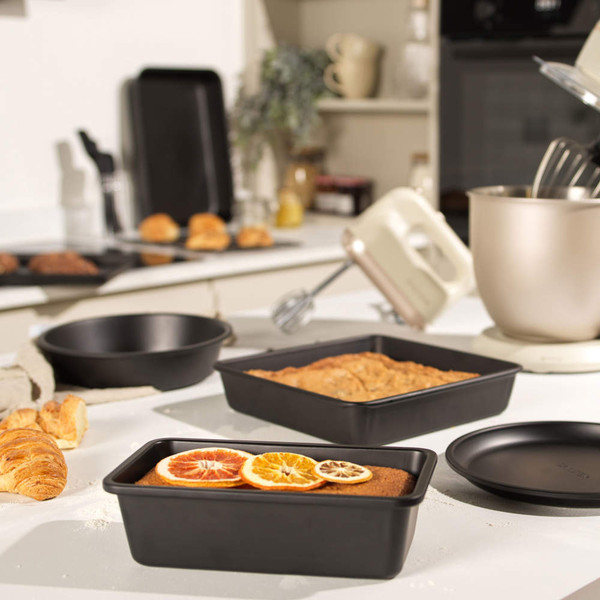 Non-stick black baking tins set with cake and bread, shown on kitchen worktop.