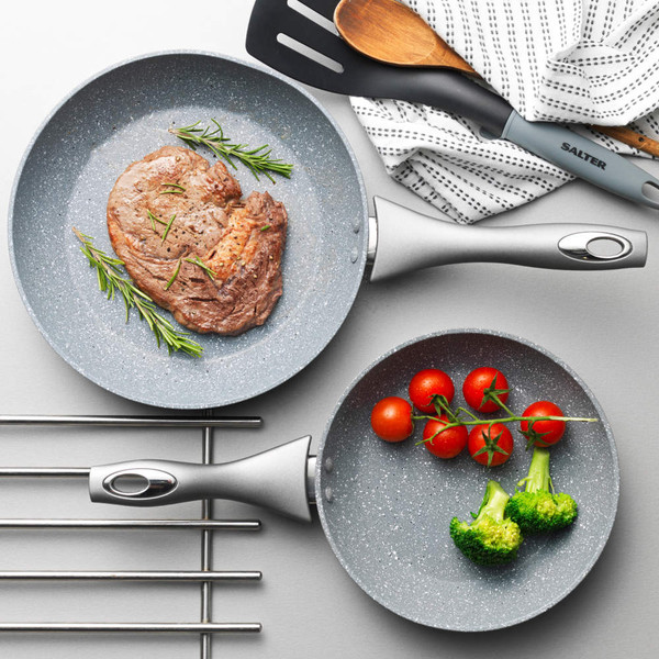 Set of two grey non-stick frying pans with steak, fresh rosemary, tomatoes, and broccoli.