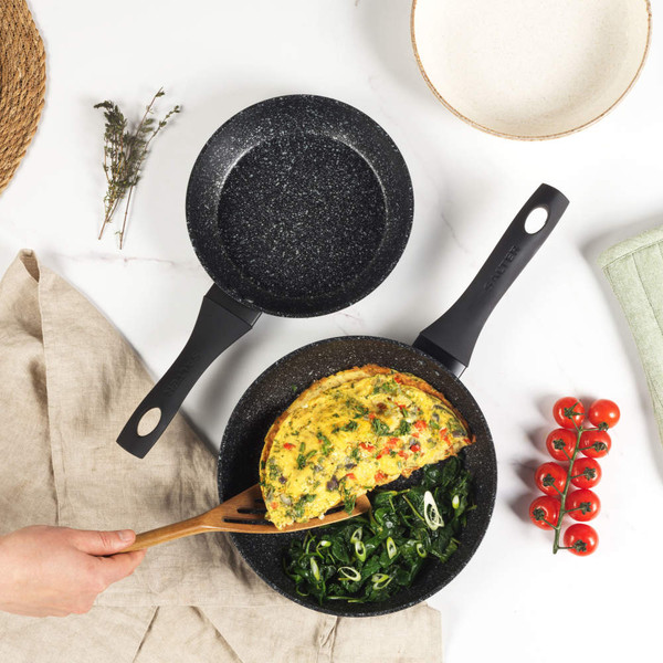 Set of two black speckled frying pans, one with omelette and greens, on kitchen counter