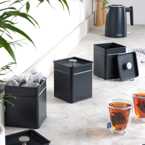 Black metal tea caddies with lids, loose tea bags and two cups of tea on kitchen counter