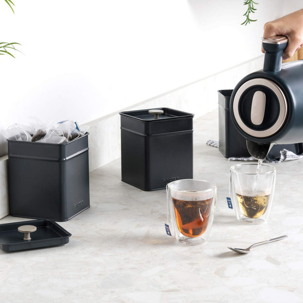 Set of black tea and coffee storage canisters with clear tea glasses on kitchen counter