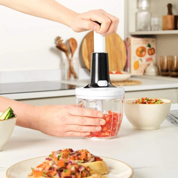 Handheld food chopper in use, chopping tomatoes in a clear container on kitchen counter.