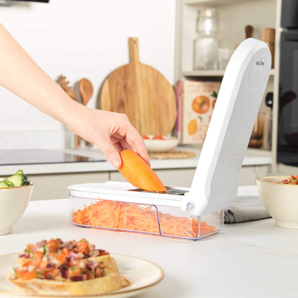 Hand using white food chopper to grate carrots into container in modern kitchen