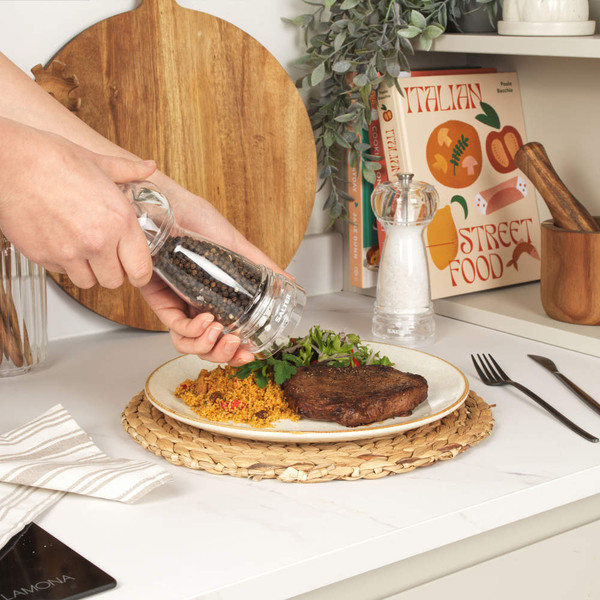 Grinding black pepper onto steak and couscous with herbs, in a kitchen setting.