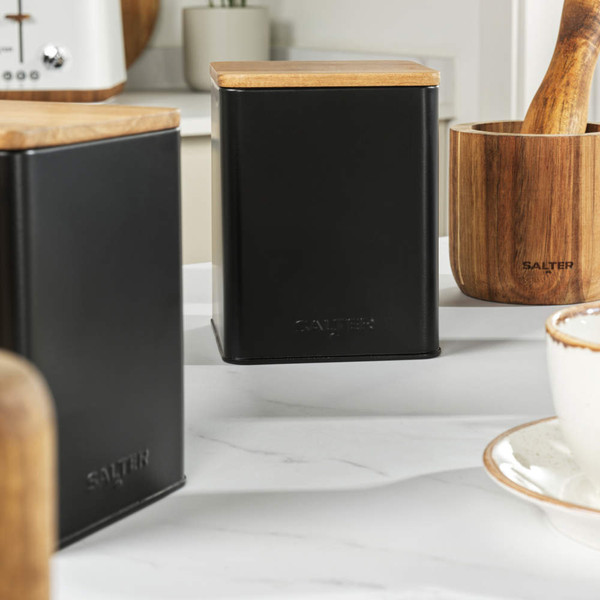 Set of three black storage canisters with wooden lids, one open with tea bags inside