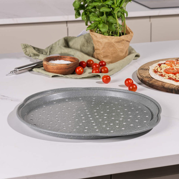 Grey non-stick pizza tray with holes, shown on kitchen counter near fresh tomatoes and herbs