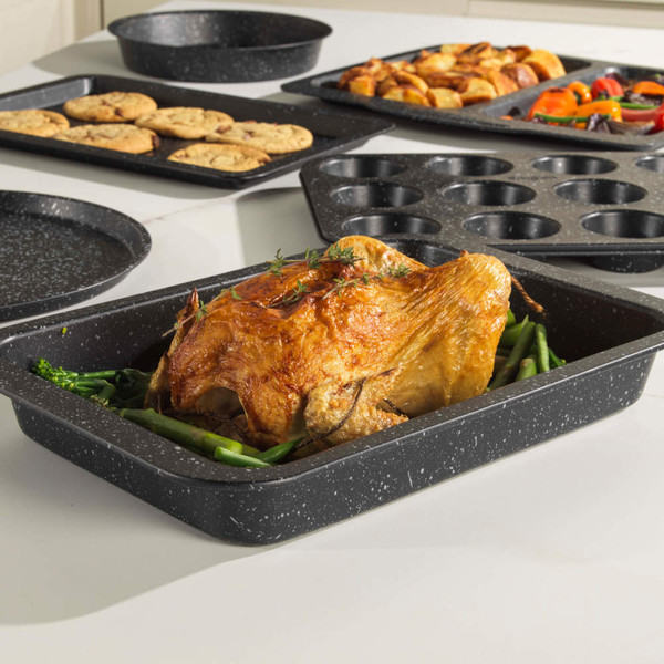 Set of non-stick black speckled bakeware, showing roasting tin with chicken, trays of cookies and vegetables.