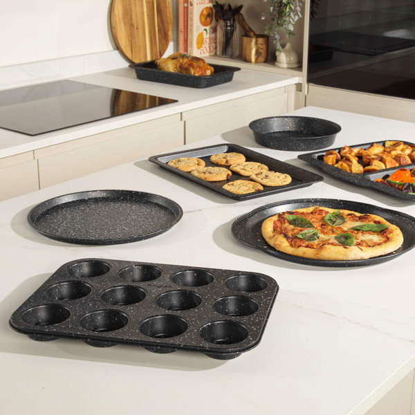 Set of black speckled non-stick bakeware on kitchen counter, includes pizza tray, roasting tray, muffin tin and cake tin.