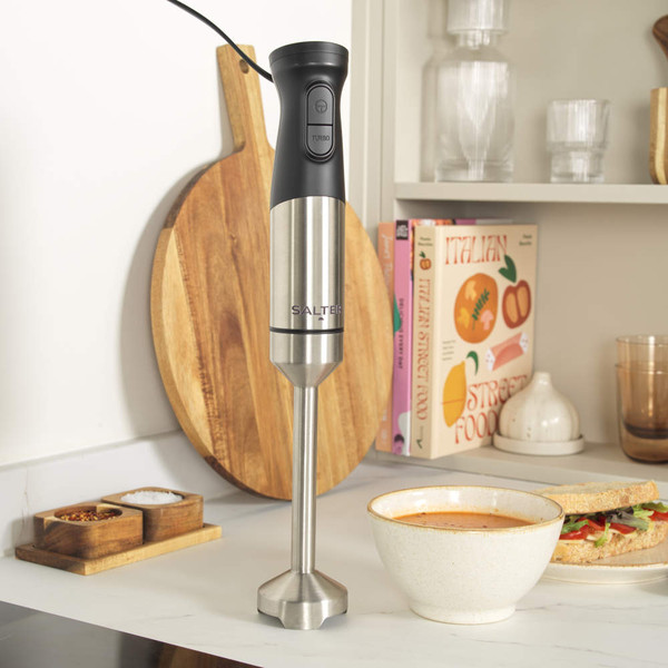 Black and silver hand blender upright on kitchen counter beside soup and sandwich