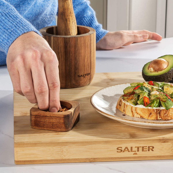 Salter wooden pestle and mortar set on bamboo board with avocado toast and seasoning.