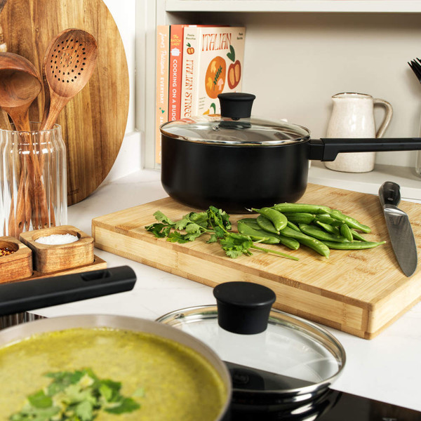 Black non-stick saucepan with glass lid on kitchen counter, beside chopped herbs and sugar snap peas